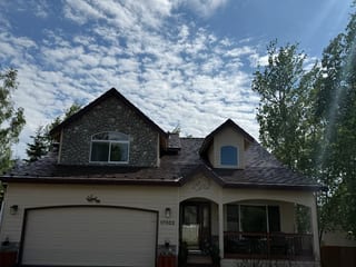A cream and brown craftsman style house with a brown metal roof and a brown garage door, set against a lush green forested backdrop under a blue sky.