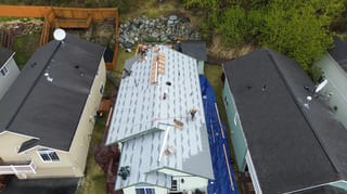 Aerial view of a ranch-style home with a gray metal roof being installed in a rural setting with lush greenery and blue skies