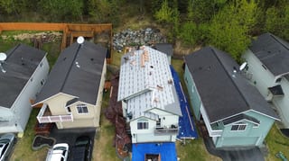 Aerial view of a white ranch-style house with a grey metal roof under construction in a forested rural area.