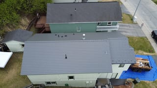 A metal roof under construction on a ranch-style home in a suburban neighborhood with lush greenery and blue sky