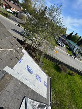 A modern single-family home with a metal roof under construction in a rural or suburban area with lush greenery and blue sky.