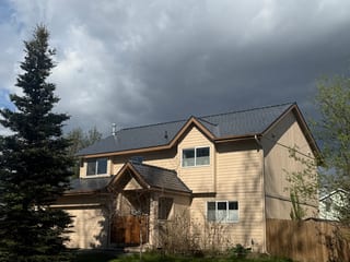 A modern-style home with a metal roof, tan siding and a brown door, under an overcast sky.