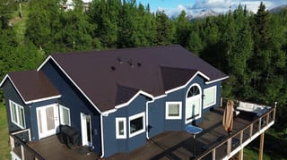 A modern single-family home with a metal roof and deck, surrounded by lush greenery and a mountain in the background.