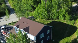 A two-story ranch-style home with a brown metal roof, white trim, and a brown deck, set against a lush green forest backdrop.