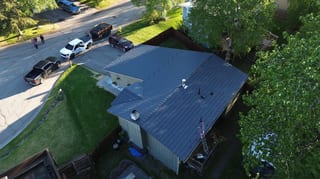Aerial view of a grey metal roof on a ranch-style home in a rural area with lush greenery and a golden hour sky.