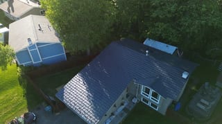 Aerial view of a completed metal roof on a ranch-style home in a wooded area with lush greenery and blue sky.