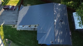 Aerial view of a dark grey metal roof on a ranch-style home in a forested area with lush greenery and blue sky.