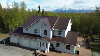 A two-story house with a brown metal roof and white siding, set amidst lush greenery and a mountain backdrop.