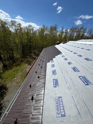 A metal roof with underlayment and Interlock detail, installed on a modern single-family home in a forested suburban area with lush greenery and a blue sky.