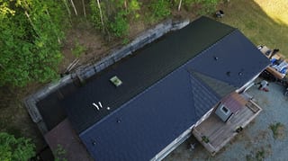 Aerial view of a modern, single-family home with a dark metal roof and solar panels in a forested rural area.