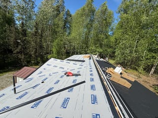 A metal roof under construction in a forested area with lush greenery and blue sky