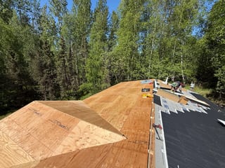 A modern-style house with a metal roof under construction, surrounded by lush greenery and a clear blue sky.