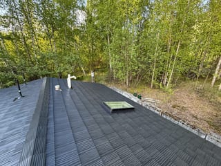 A modern single-family home with a metal roof, set in a forested area with lush greenery and a clear blue sky.