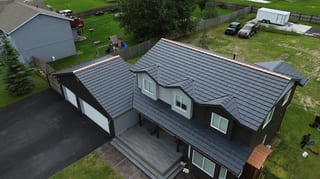 Aerial view of a modern single-family home with a dark grey metal roof and white trim, situated in a suburban neighborhood with lush greenery and blue skies.