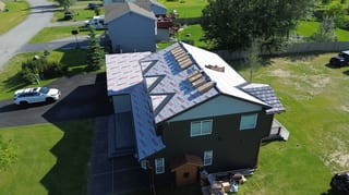 Aerial view of a modern style single-family home in a suburban area with a metal roof under construction, featuring a chimney, ridge cap, ridge vent, snow guard