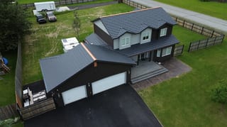 Aerial view of a modern single-family home with a black metal roof and white garage doors, set in an urban setting with lush greenery and blue sky.
