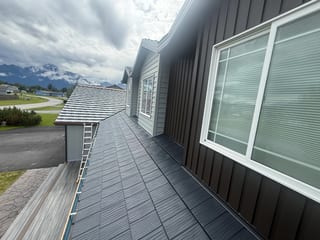 A modern home with a dark grey metal roof, set against a lush green mountain backdrop under an overcast sky.