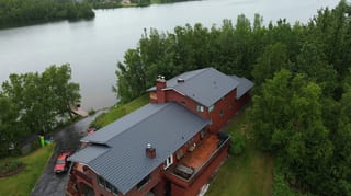 A ranch-style home with a red brick exterior and a grey metal roof, set amidst lush greenery and overlooking a serene body of water.