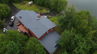 Aerial view of a red brick house with a grey metal roof, surrounded by lush green trees.