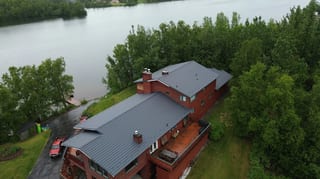 Aerial view of a large, red, modern house with a grey metal roof, situated on the waterfront, surrounded by lush greenery on a cloudy day.