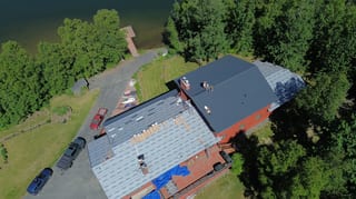 Metal roof installation in progress on a large, red, brick house with grey shingles on a sunny day.