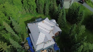 A colonial-style home with a white metal roof and a chimney, surrounded by lush greenery