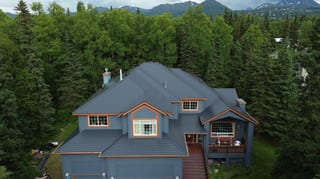 A modern single-family home with a grey metal roof and brown trim, set against a lush forested backdrop with a mountain range in the distance.