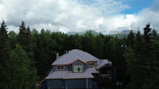 Aerial view of a finished metal roof on a chalet-style home in a forested mountain setting, with lush greenery and blue sky
