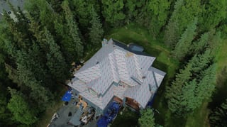 A modern single-family home with a grey metal roof and white trim, surrounded by lush greenery and set in a rural forested area.