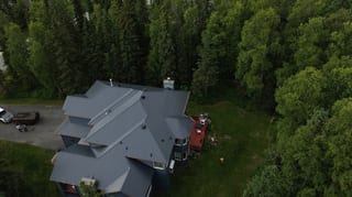 Aerial view of a modern single-family home with a grey metal roof, surrounded by lush greenery in a forested rural area.
