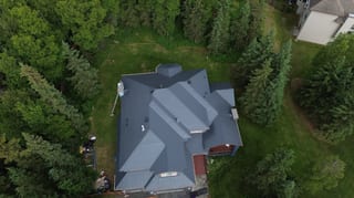 Aerial view of a modern single-family home with a grey metal roof, surrounded by lush greenery and set in a rural forested area.