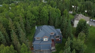 A large Colonial-style home with a grey metal roof and a chimney, set in a forested area surrounded by lush greenery and a clear blue sky.