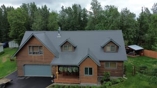 A cottage with a grey metal roof and brown siding, surrounded by lush greenery and trees.