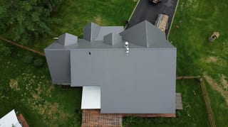 Aerial view of a modern, single-family home with a grey metal roof, snow guards and rails, and a roof louver, in a rural setting with lush greenery.