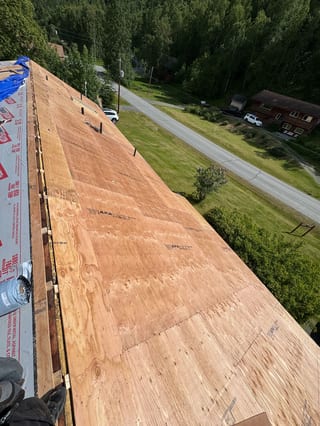 A modern-style home with a metal roof under construction, set amidst a lush forested area with a rural backdrop.
