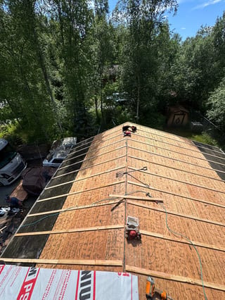 A modern-style metal roof being installed on a single-family home in a forested area with lush greenery and a blue sky.