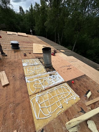 A colonial-style single-family home in a forested rural area with lush greenery and overcast skies, during installation of a metal roof.