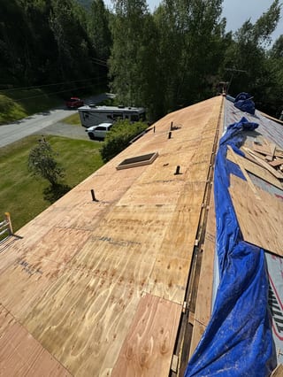 A partially installed metal roof on a ranch-style single-family home in a forested rural setting with a blue sky.