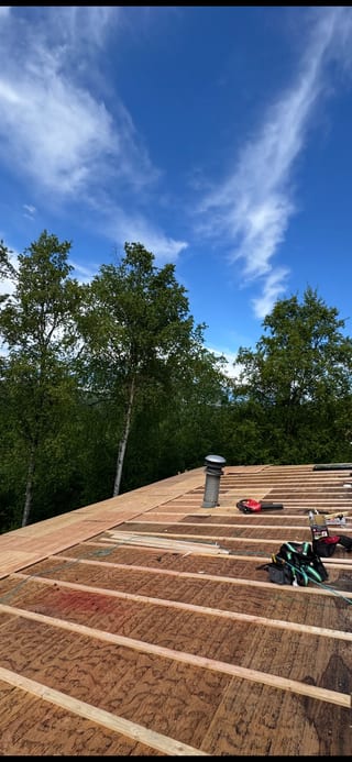 A metal roof under construction in a forested area with lush greenery and a blue sky.