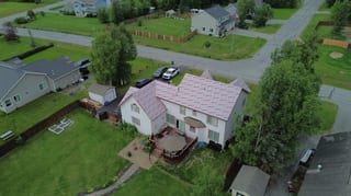 Aerial view of a colonial-style home with a pink metal roof in a rural setting, surrounded by lush greenery and a blue sky