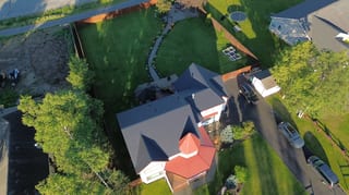 A colonial-style home with a dark grey metal roof and red trim, surrounded by lush greenery and a blue sky.