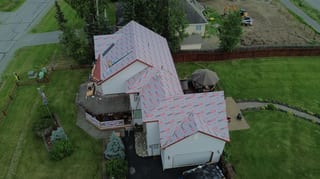 Aerial view of a metal roof being installed on a ranch-style single-family home in a suburban area with lush greenery.