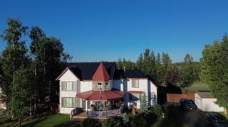 A colonial-style home with a red metal roof and white siding, set amidst lush greenery under a clear blue sky.