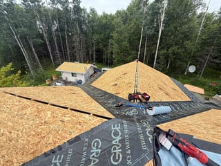 A modern single-family home with a metal roof under construction in a forested rural setting.
