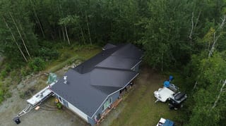 A metal roof with a dark grey colour on a single-family ranch-style home in a forested setting, with lush greenery and overcast conditions.