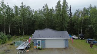 A ranch-style home with a metal roof and lush greenery, located in a forested rural area.