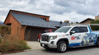 Slate-blue Interlock Steel Slate metal roof on a 1.5-storey Craftsman home in Vernon BC, viewed from the front-left with an Interlock GMC Sierra service truck in foreground.