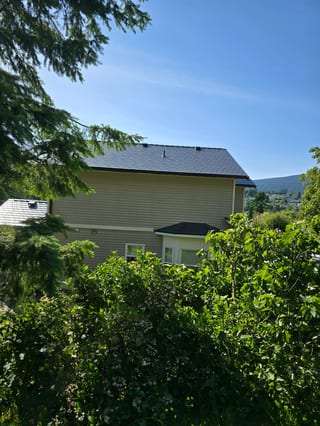 Deep Charcoal Interlock Slate aluminum roof on a tan two-storey home with dormers among tall pines, Gibsons, British Columbia