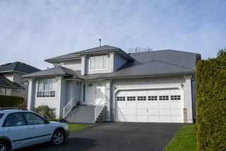 Musket Grey Interlock Slate aluminum roof on a white two-storey home with dormers among tall pines, Surrey, British Columbia