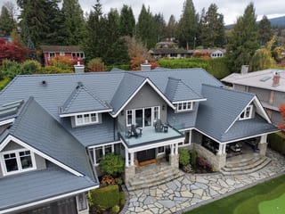 Deep Charcoal Interlock Slate aluminum roof on a gray two-storey home with dormers among tall pines, North Vancouver, British Columbia
